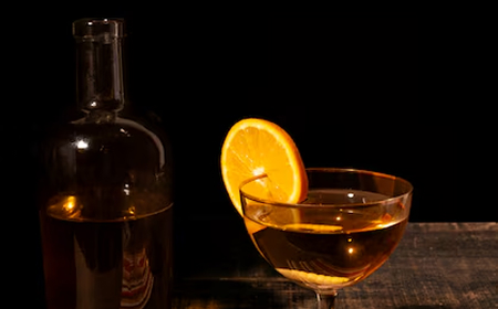 Бутылка и бокал с янтарным напитком, украшенным долькой апельсина, на тёмном фоне